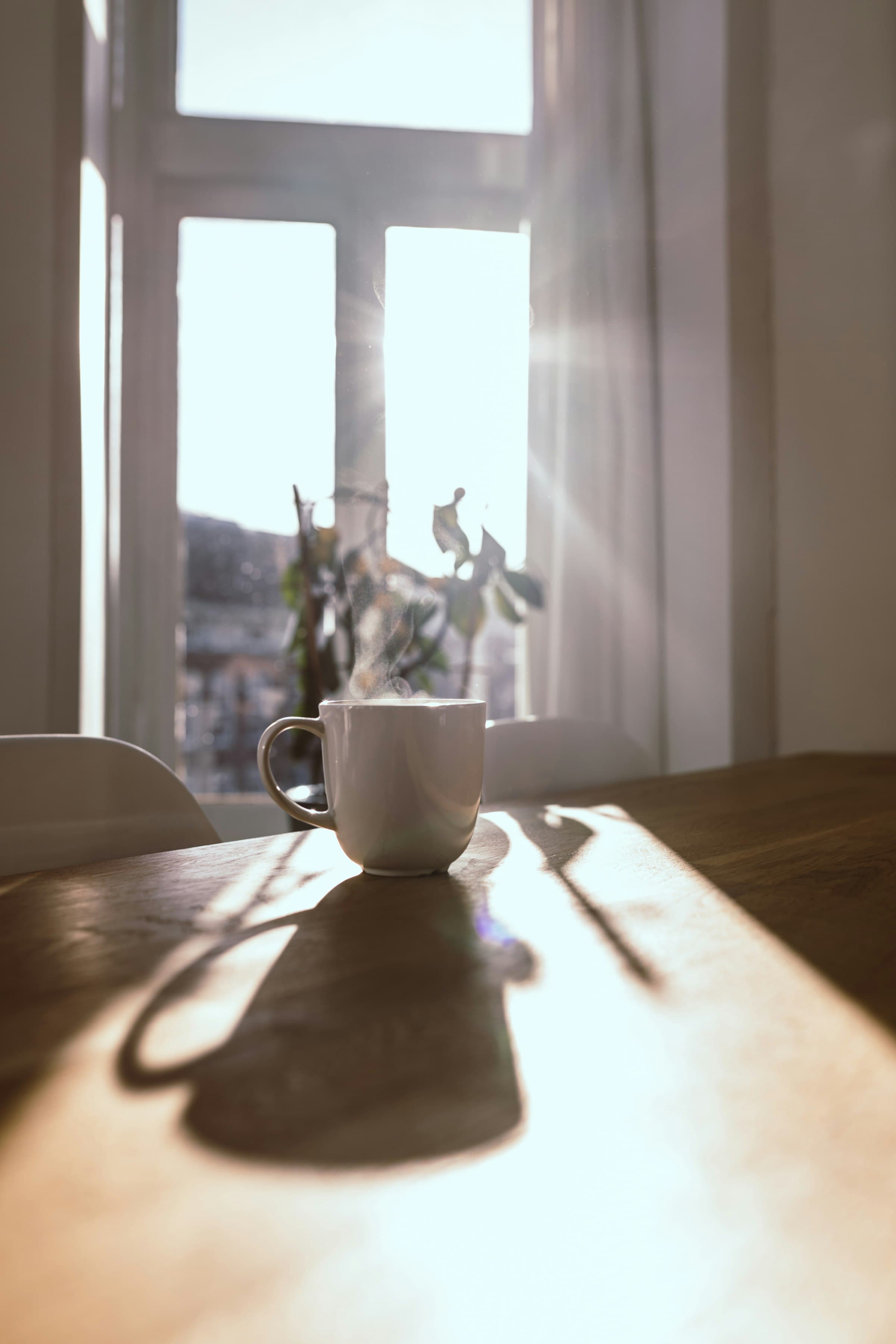 A steaming white mug on a wooden table in front of a sunlit window.