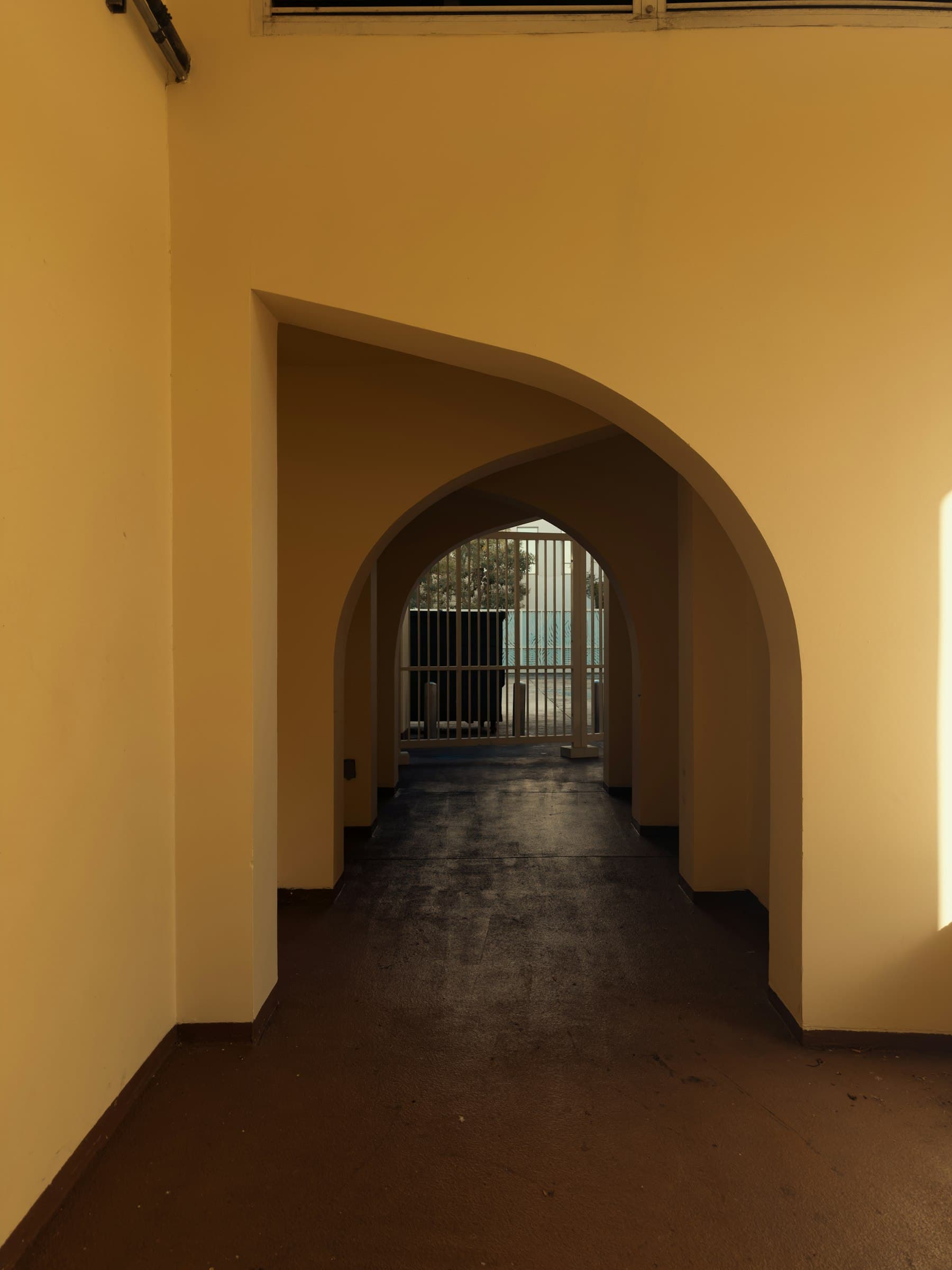 A corridor of ochre plaster archways leading to a barred gate and daylight.