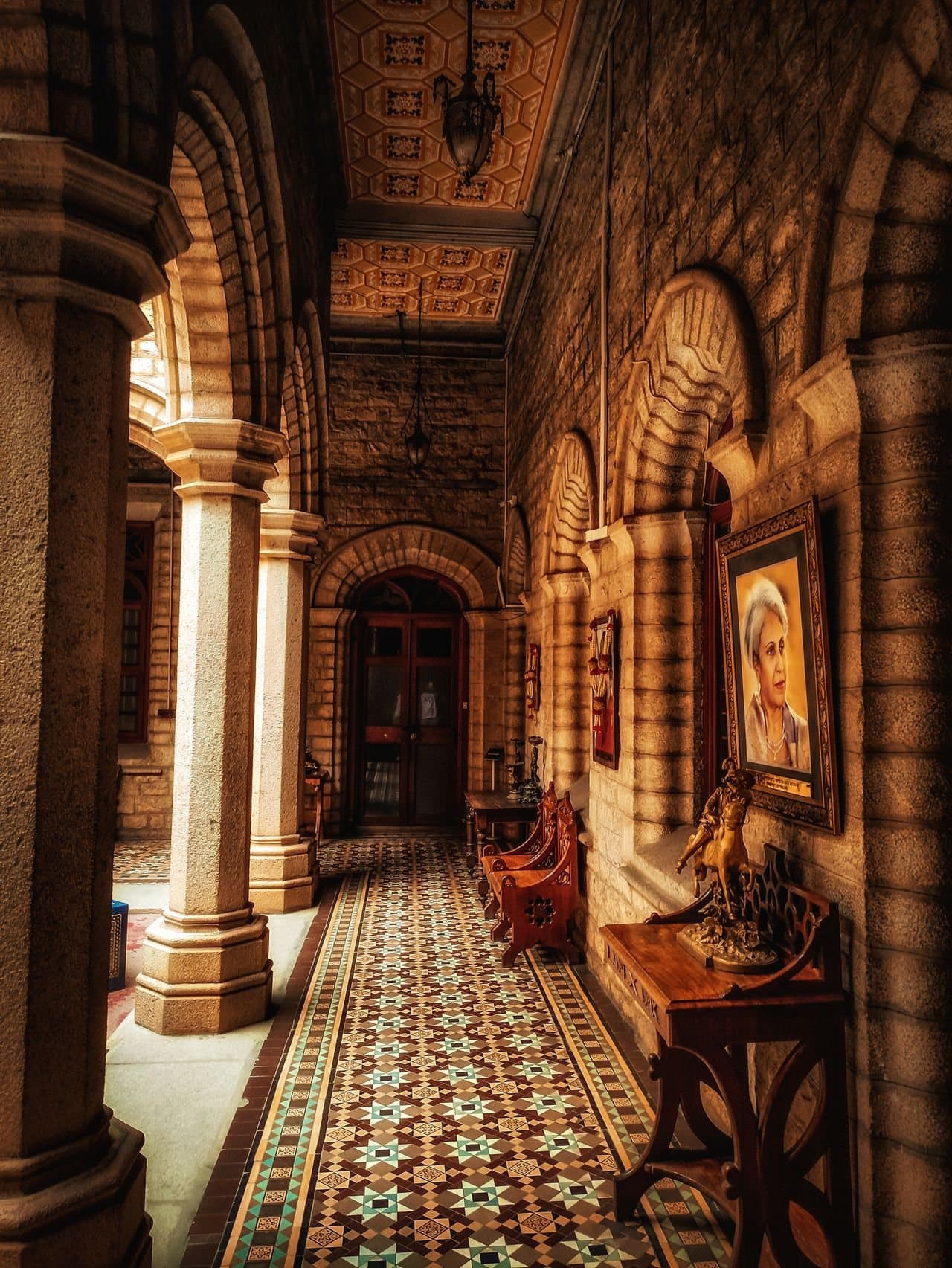 A stone colonnade inside Bangalore Palace, with arched walls, a patterned tiled floor and carved wooden benches.