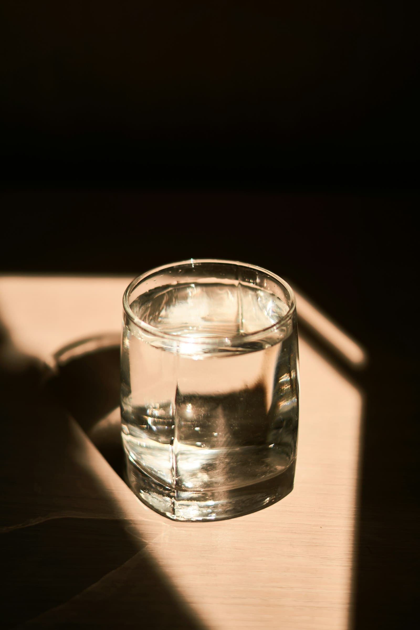 A glass of water on a table catching a shaft of warm sunlight between dark shadows.